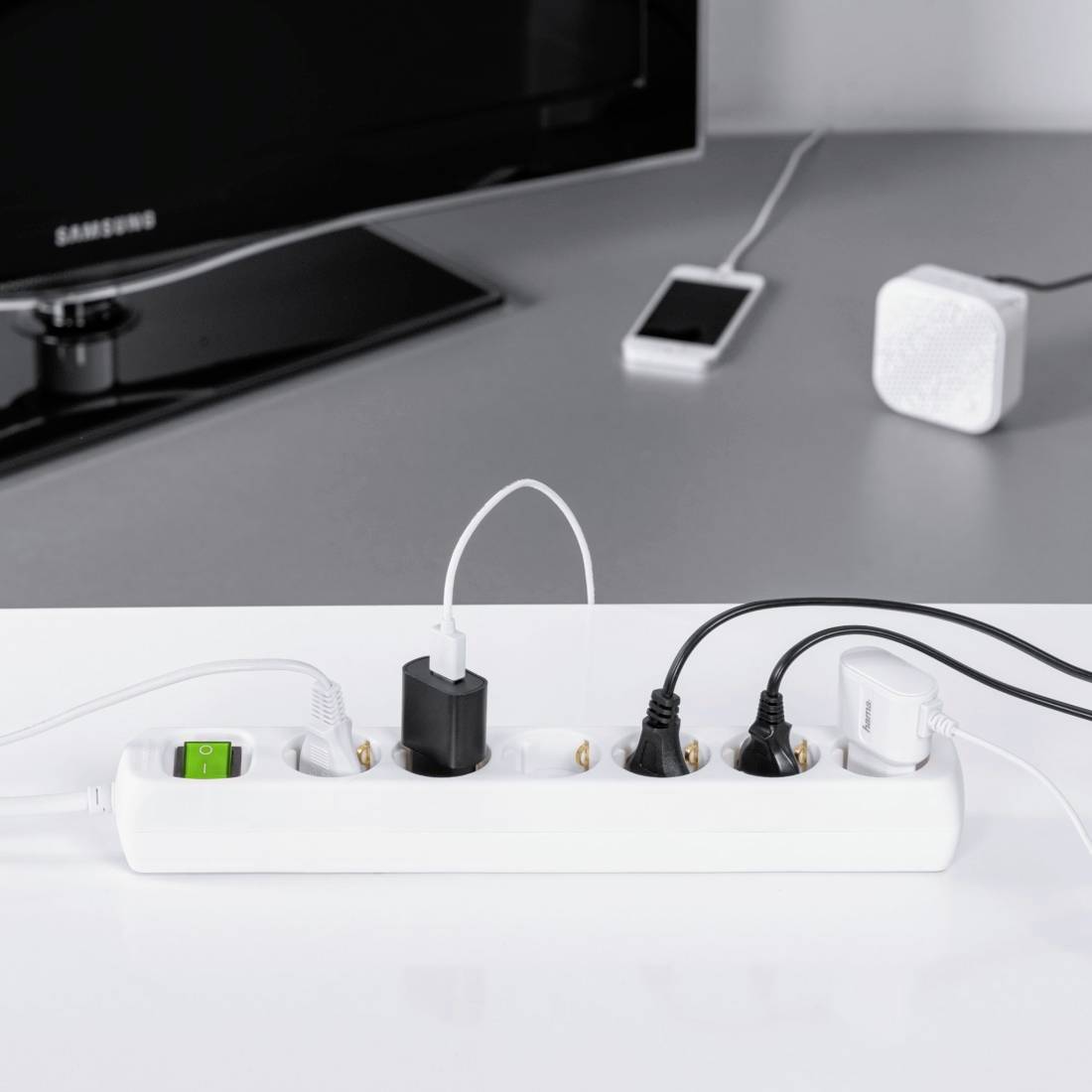 Eine Steckdosenleiste auf einem Tisch versorgt elektronische Geräte wie ein Smartphone und Lautsprecher. Ein TV steht im Hintergrund.