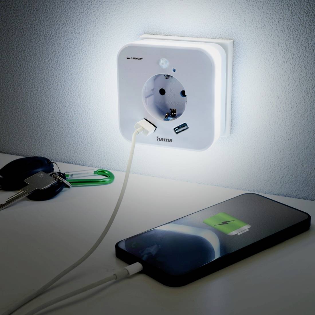Ein Smartphone wird auf einem Tisch aufgeladen. Es ist an eine leuchtende Steckdose in der Wand angeschlossen, neben einem Schlüsselbund.