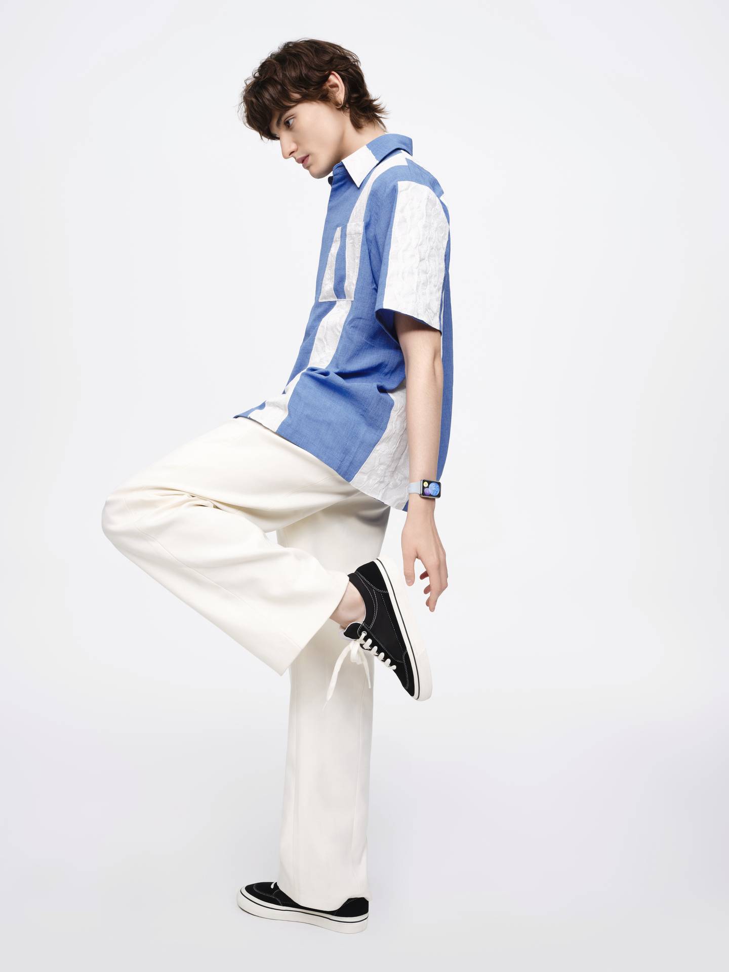 Eine Person im Profil trägt ein blau-weiß gestreiftes Hemd und weiße Hosen, hebt ein Bein an und trägt schwarze Sneakers. Minimalistischer Hintergrund.