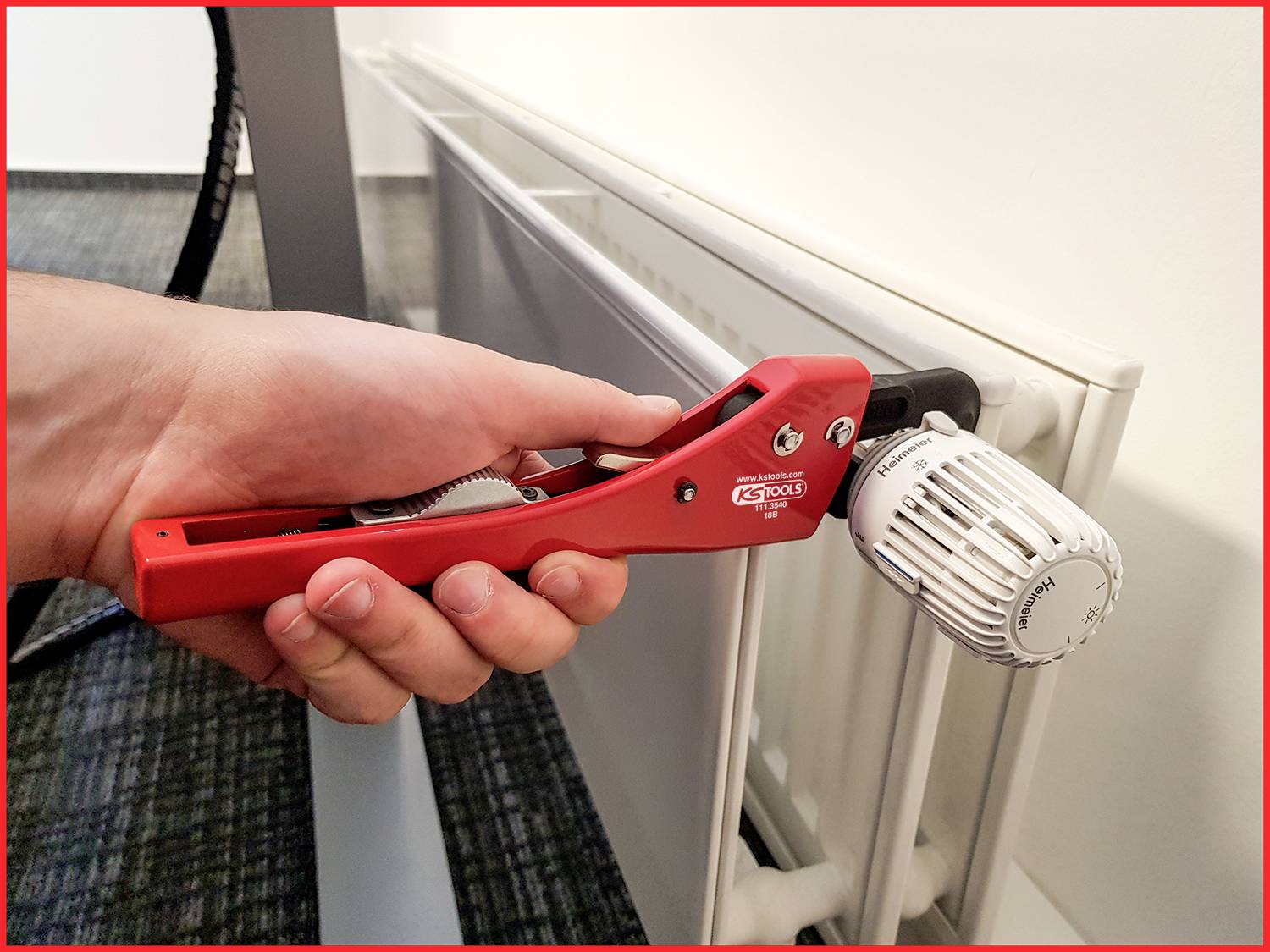 Eine Hand hält ein rotes Werkzeug und dreht vorsichtig das Thermostatventil eines weißen Heizkörpers in einem Büro.