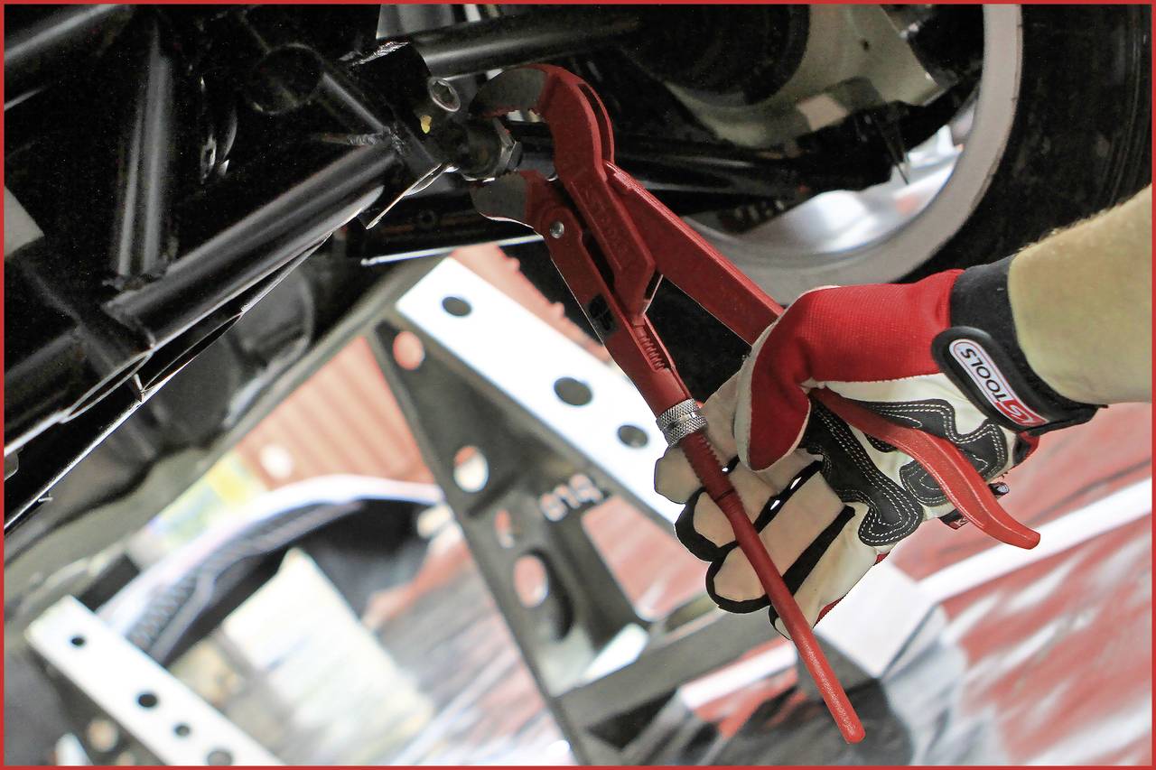 Ein Werkzeug wird von einer Person mit Handschuh an einem Auto unterhalb des Fahrzeugs verwendet, vermutlich zur Wartung oder Reparatur.