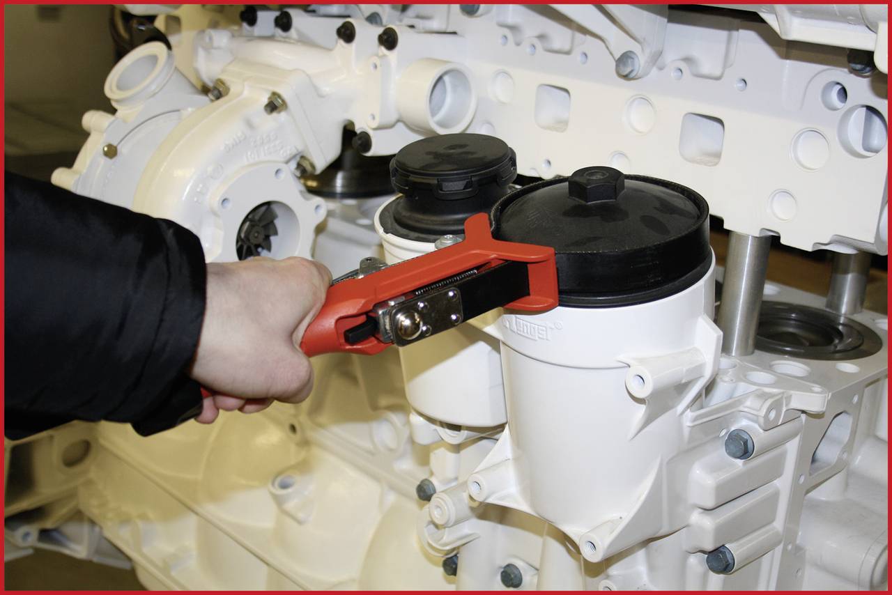 Eine Person benutzt ein rotes Werkzeug, um den schwarzen Deckel eines weißen Filters an einer großen Maschine zu drehen oder zu öffnen.