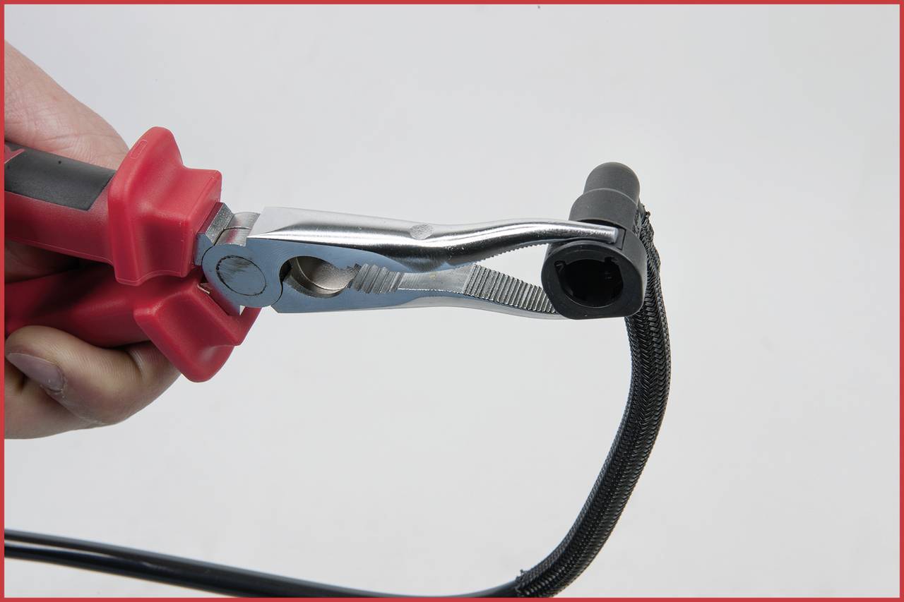 Eine Hand hält ein rotes Werkzeug, das eine Klemme zusammendrückt. Ein schwarzes Kabel ist zwischen den Klemmen eingespannt.