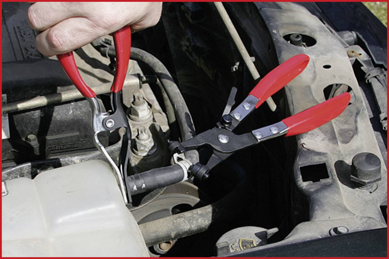 Eine Person verwendet zwei rote Zangen, um an einem Autoteil zu arbeiten. Der Motorraum und Schläuche sind sichtbar.