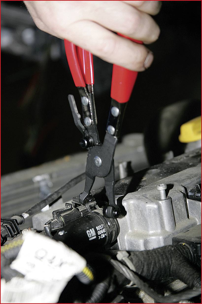 Hand hält rote Zange, greift an einem schwarzen Bauteil auf einem Motorblock. Mechanische Arbeit an einem Fahrzeugmotor.
