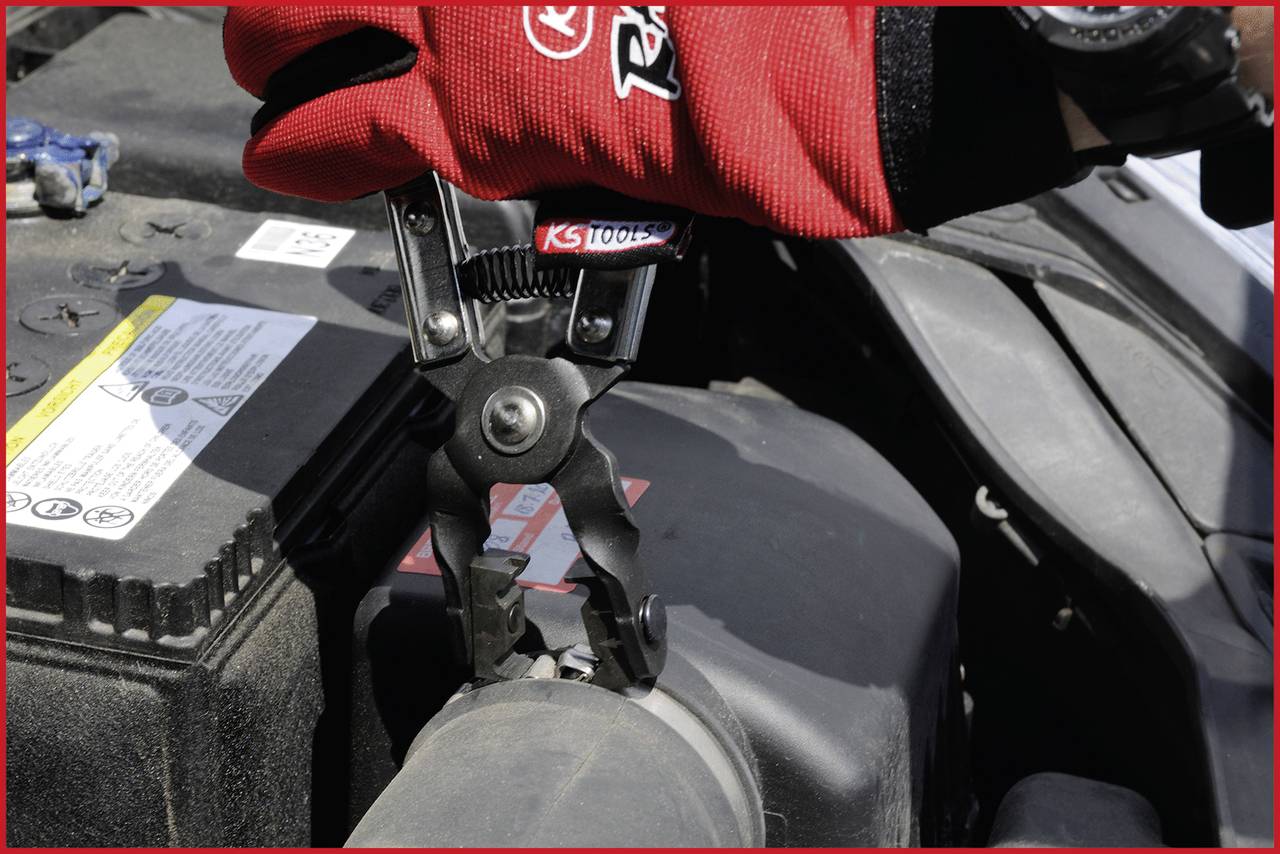 Linke Hand mit rotem Handschuh verwendet Zange, um Schlauchklammer im Motorraum eines Fahrzeugs zu lösen. Batterie im Hintergrund.