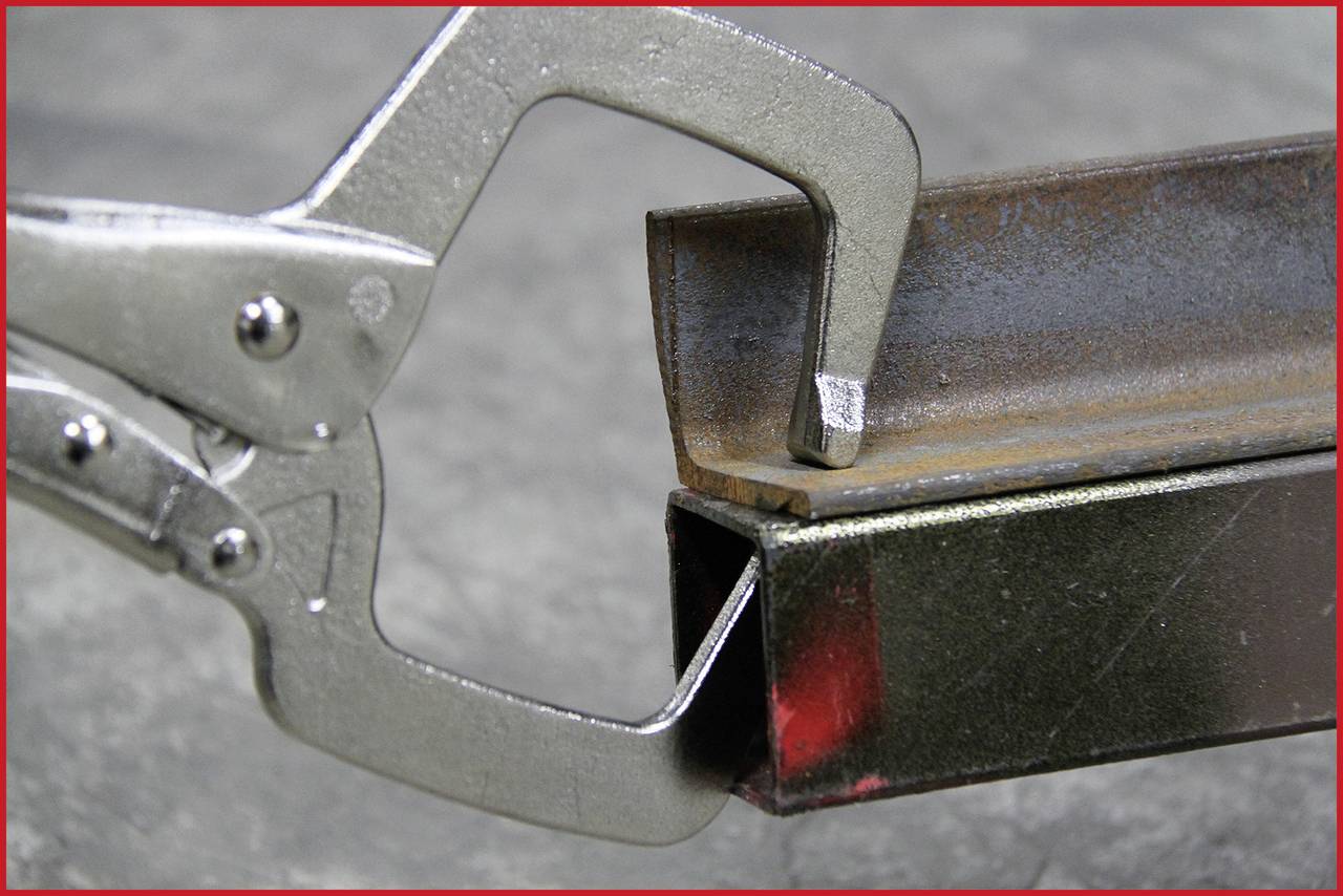 Ein Metallgreifer hält ein L-Profil gegen ein Vierkantrohr. Beide Gegenstände aus Metall. Industrialle Umgebung.