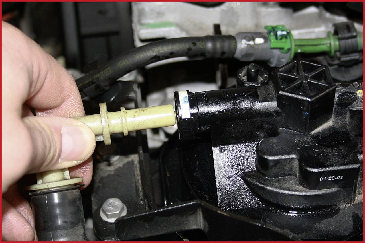 Eine Hand hält einen gelben Plastikschlauch und steckt ihn in ein schwarzes mechanisches Bauteil in einem Motorraum.