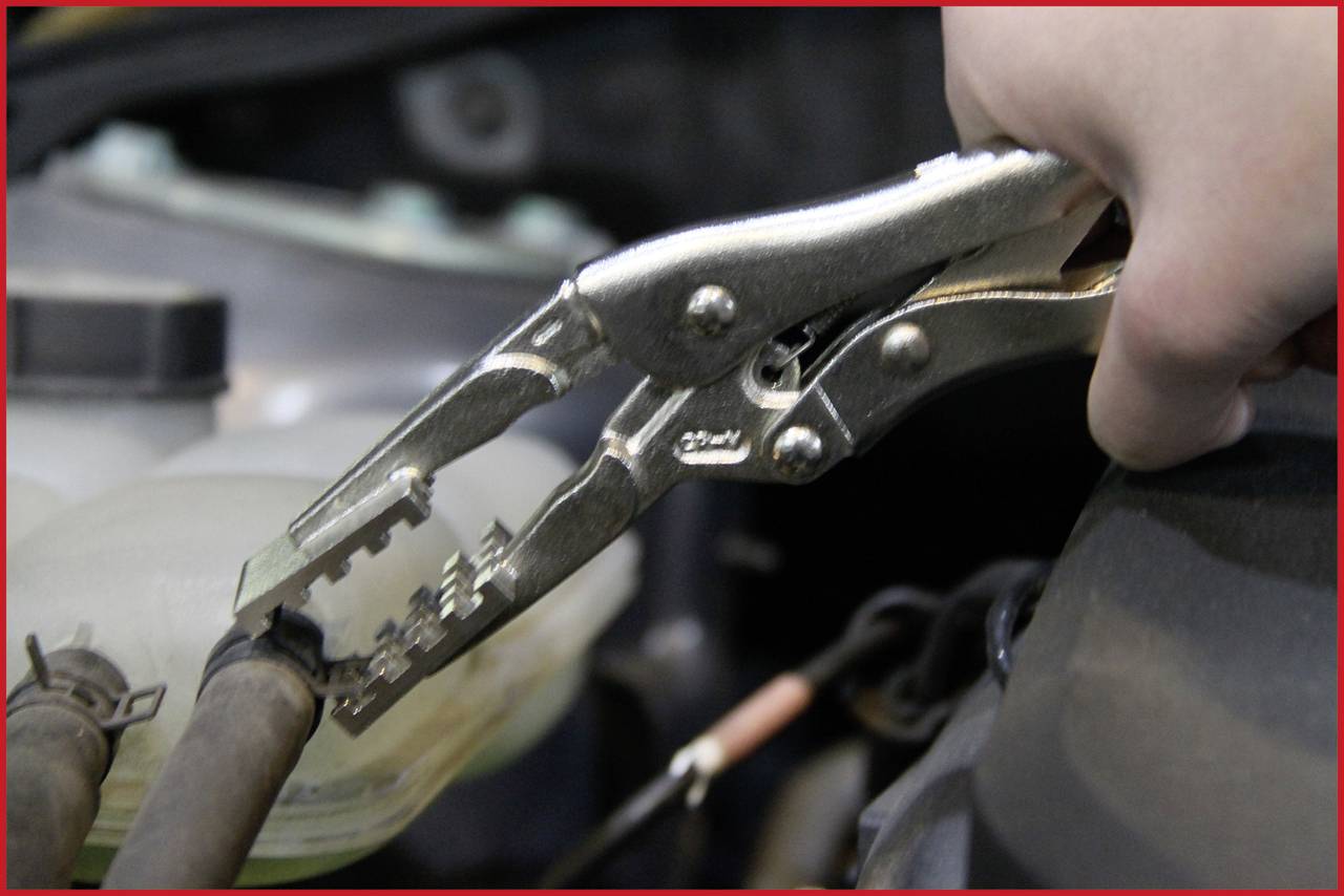Eine Hand hält eine Rohrzange, die an einem Schlauch im Motorraum eines Fahrzeugs befestigt ist, offenbar für Reparaturarbeiten.