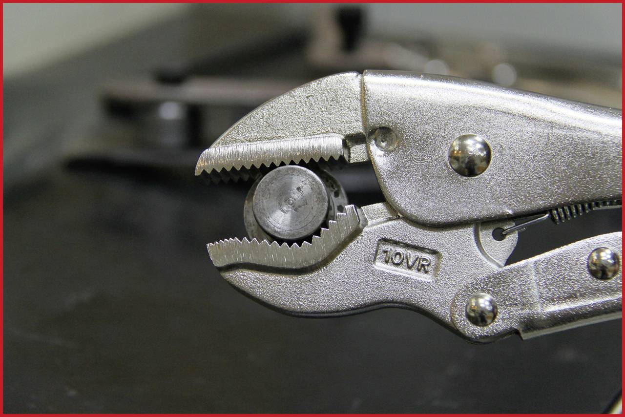 Eine silberne Zange hält eine kleine Metallmutter fest. Im Hintergrund sind verschwommene Werkzeuge zu sehen.