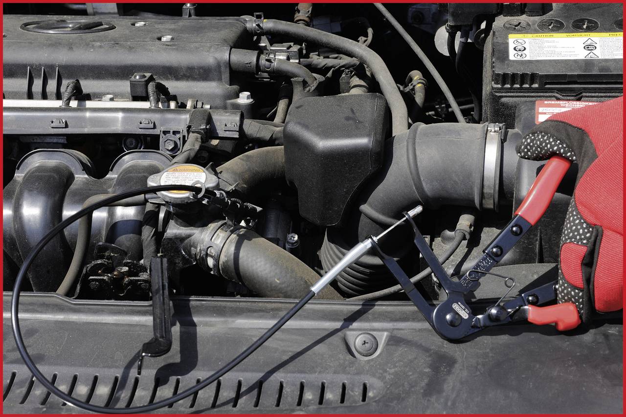 Eine Hand in einem roten Handschuh benutzt eine Zange, um einen Schlauch im Motorraum eines Autos zu klemmen.