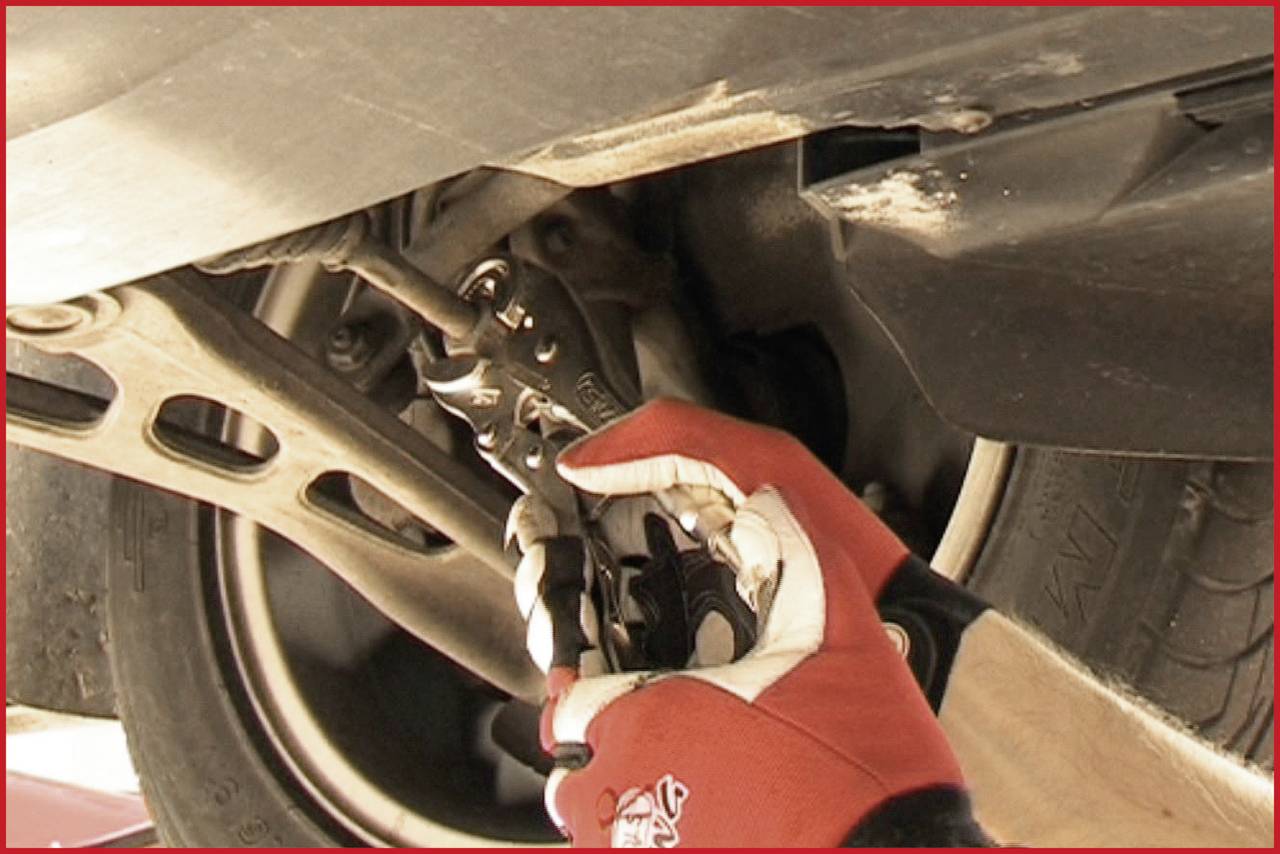Eine Person mit roten Handschuhen verwendet eine Zange, um an einem Fahrzeugteil unter dem Auto zu arbeiten. Das Auto ist aufgebockt.