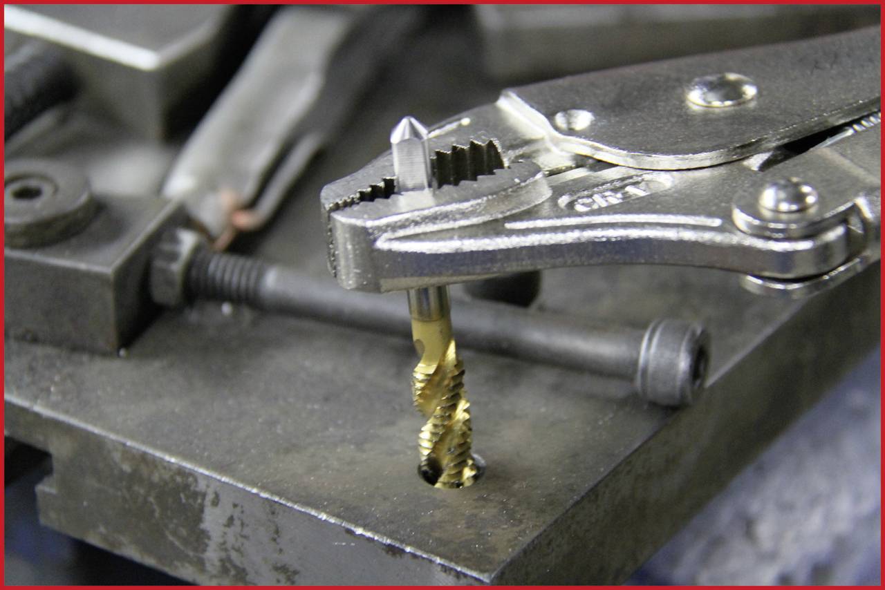 Eine Zange hält einen Gewindebohrer, der in einem Metallblock eingespannt ist. Im Hintergrund befinden sich Werkzeuge und Schrauben.
