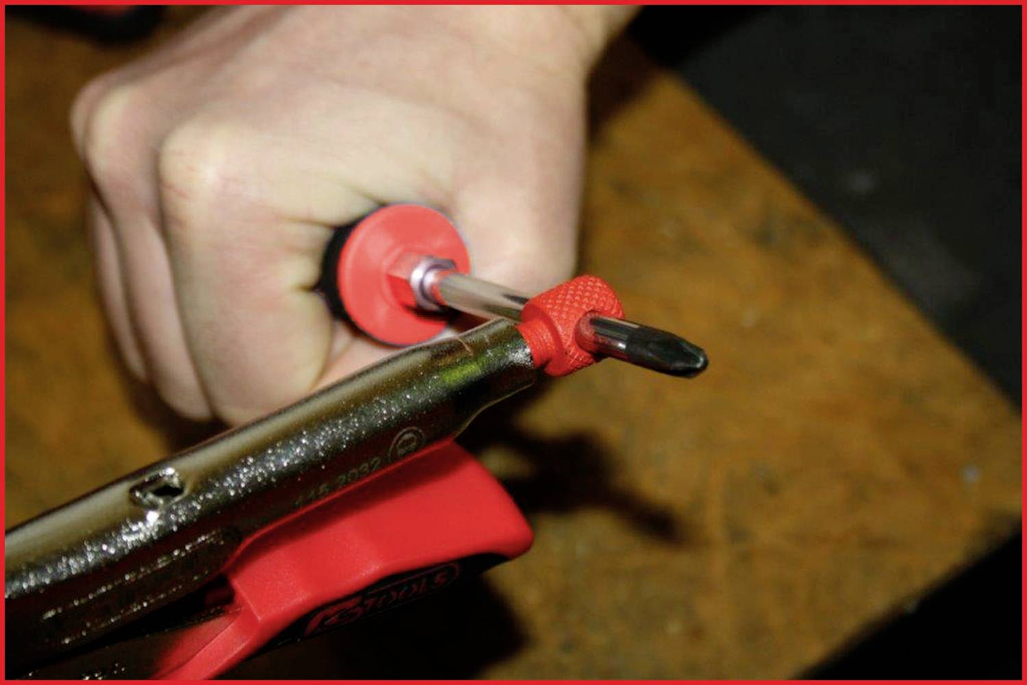Eine Person hält einen Schraubendreher mit einem roten Griff, während sie eine rote Klemme verwendet, um ihn auf einem Holztisch zu fixieren.