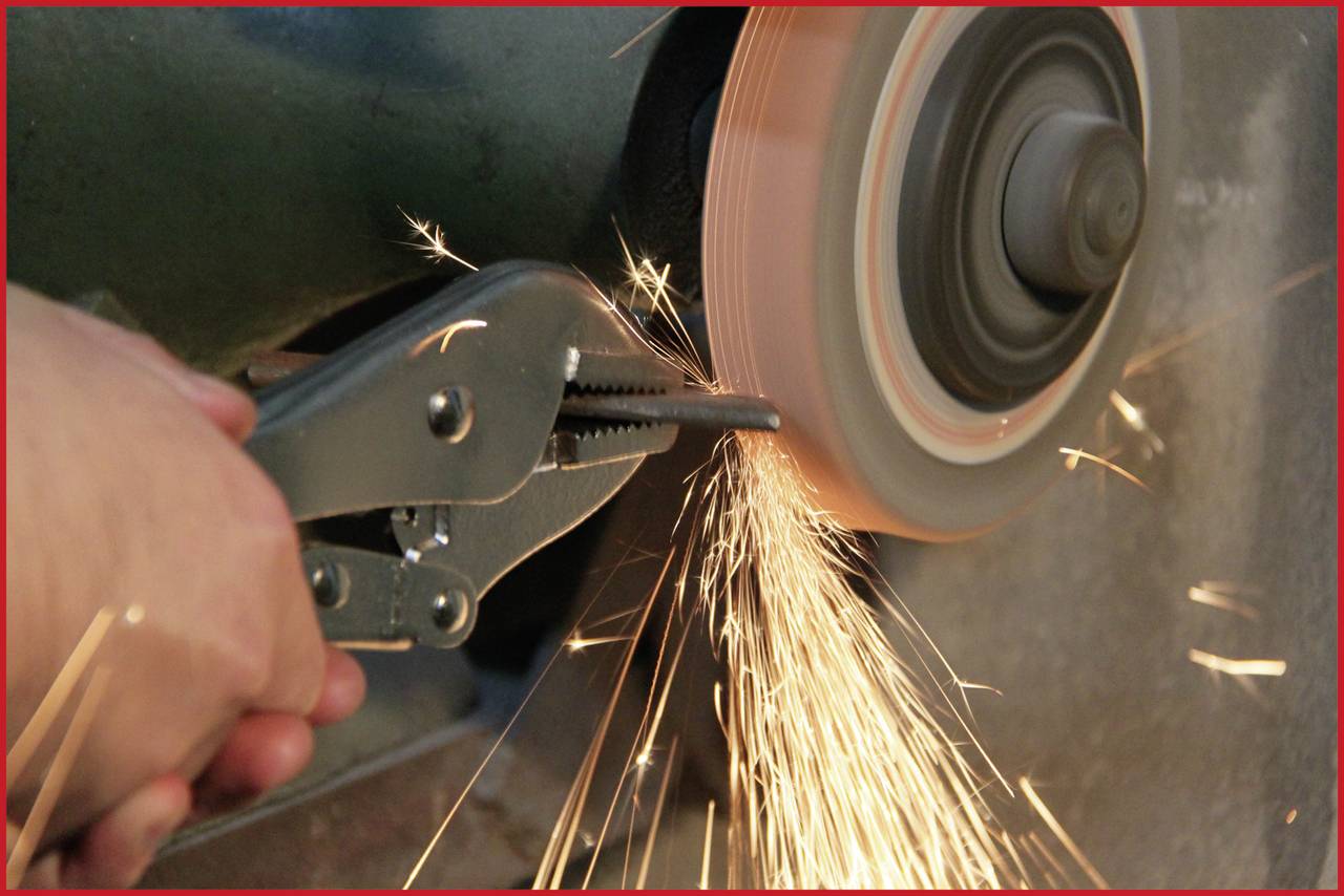 Eine Hand hält eine Zange, die ein Metallstück gegen ein rotierendes Schleifrad drückt, wobei Funken sprühen.