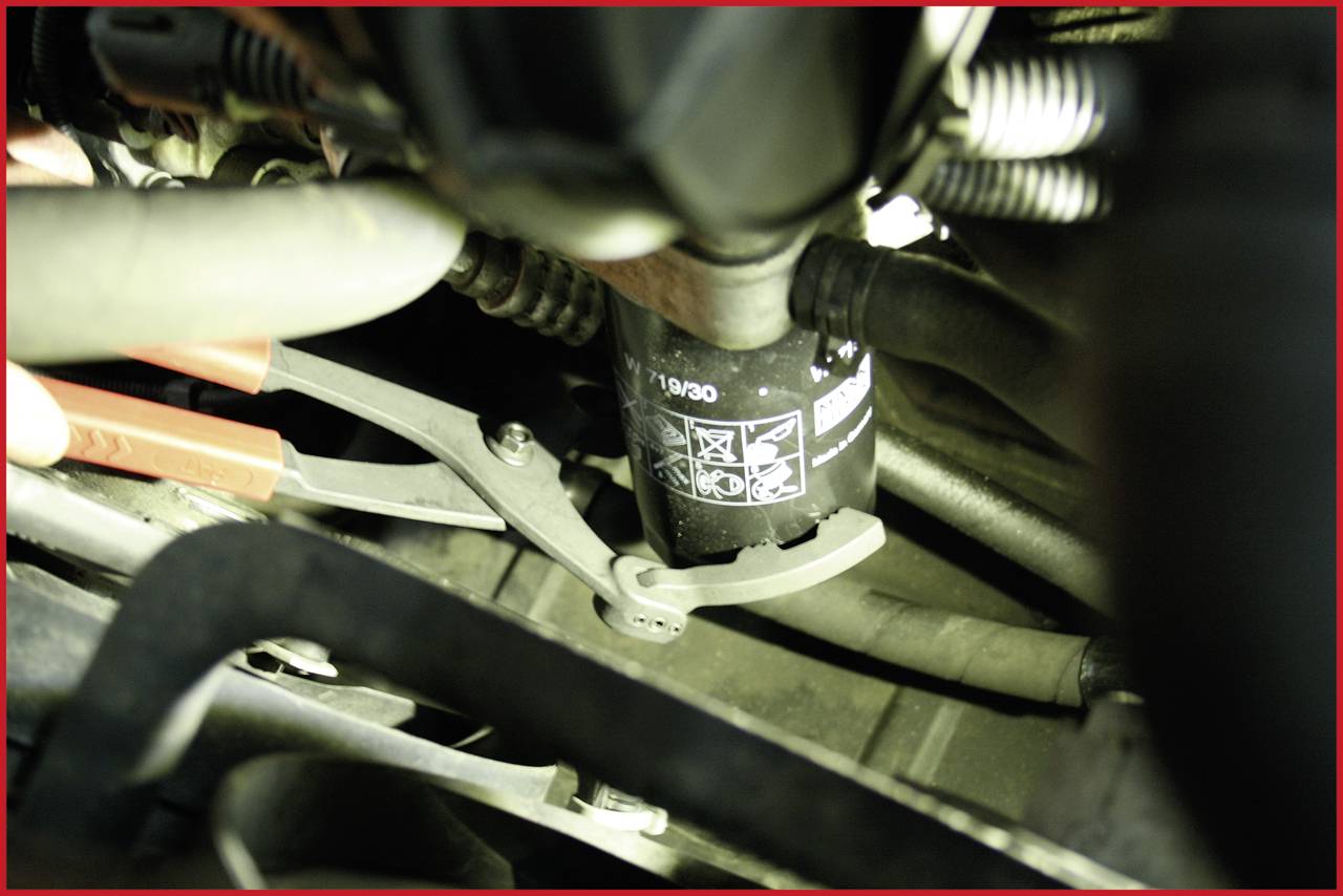 Naheaufnahme eines Ölfilters in einem Motorraum, der mit einer Rohrzange festgehalten wird. Der Filter zeigt Symbole zur Installation.