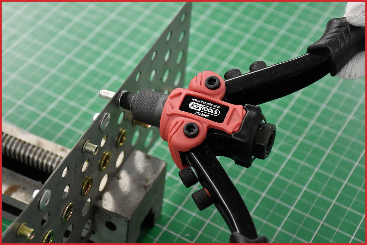 Eine rote Nietzange wird verwendet, um eine Nietmutter an ein perforiertes Blech zu befestigen. Werkzeug und Werkstück auf grünem Hintergrund.