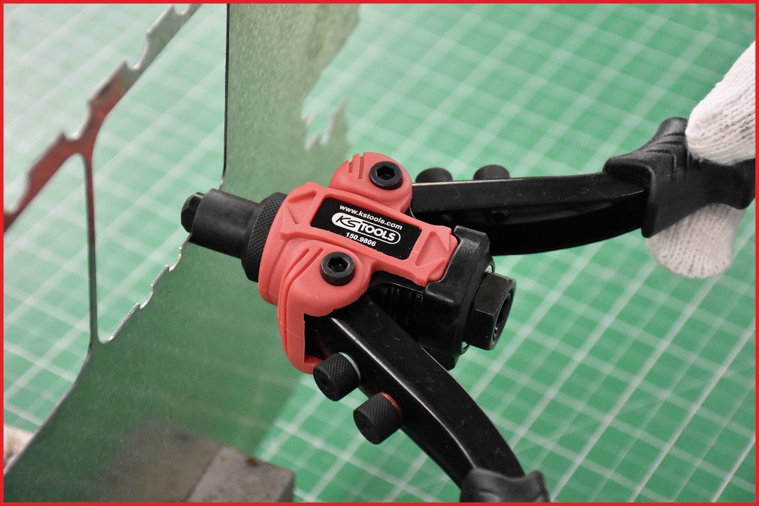 Eine Person schneidet ein Stück Blech mit einer roten Blechschere auf einem grünen Gitterhintergrund.