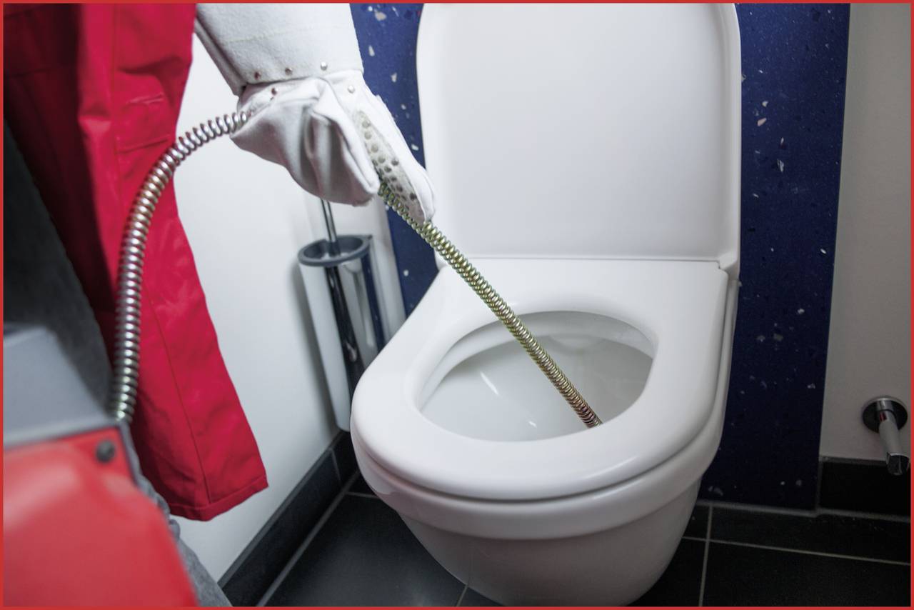 Eine Person in Schutzkleidung reinigt eine Toilette mit einer langen Reinigungsbürste.