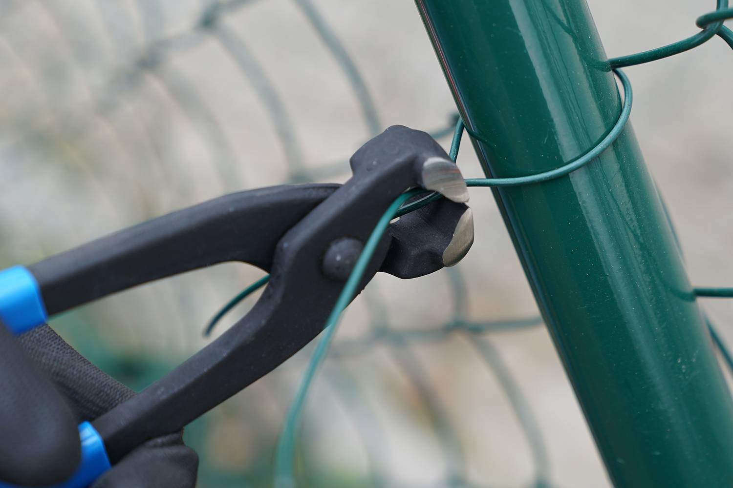 Ein Seitenschneider durchtrennt einen grünen Draht, der an einem grünen Metallzaun befestigt ist.