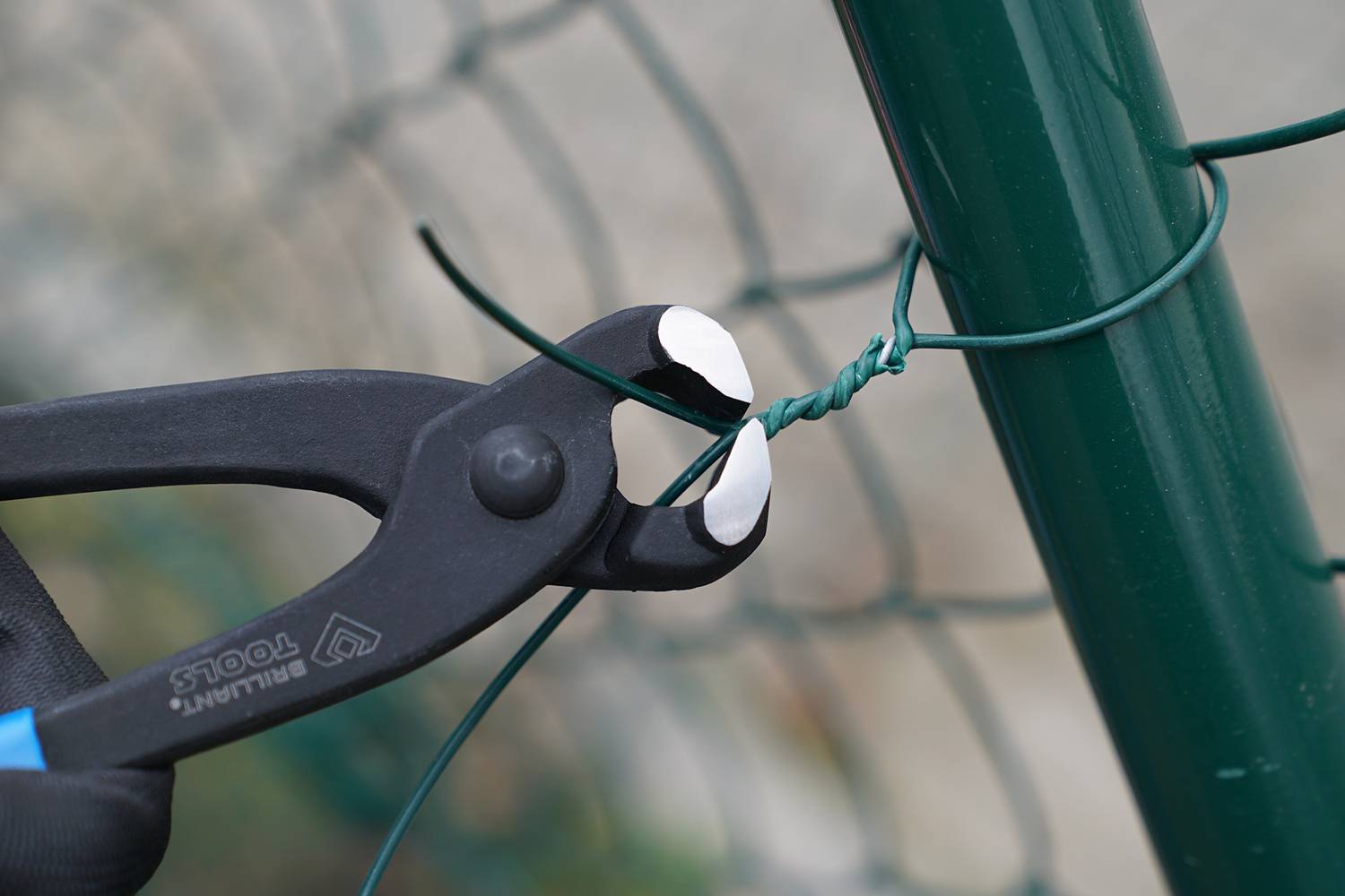 Zange schneidet Draht nahe an einem grünen Metallzaun. Eine Person hält die Zange, während der Draht durchtrennt wird.