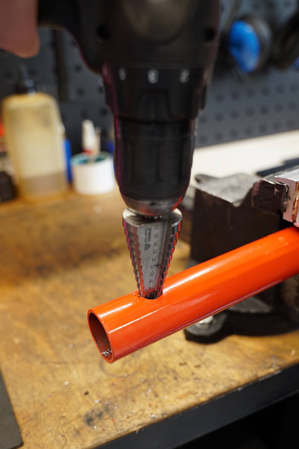 Bohrmaschine mit Kegelbohrer, bohrt ein Loch in ein orangefarbenes Metallrohr auf einer Werkbank in einer Werkstattumgebung.