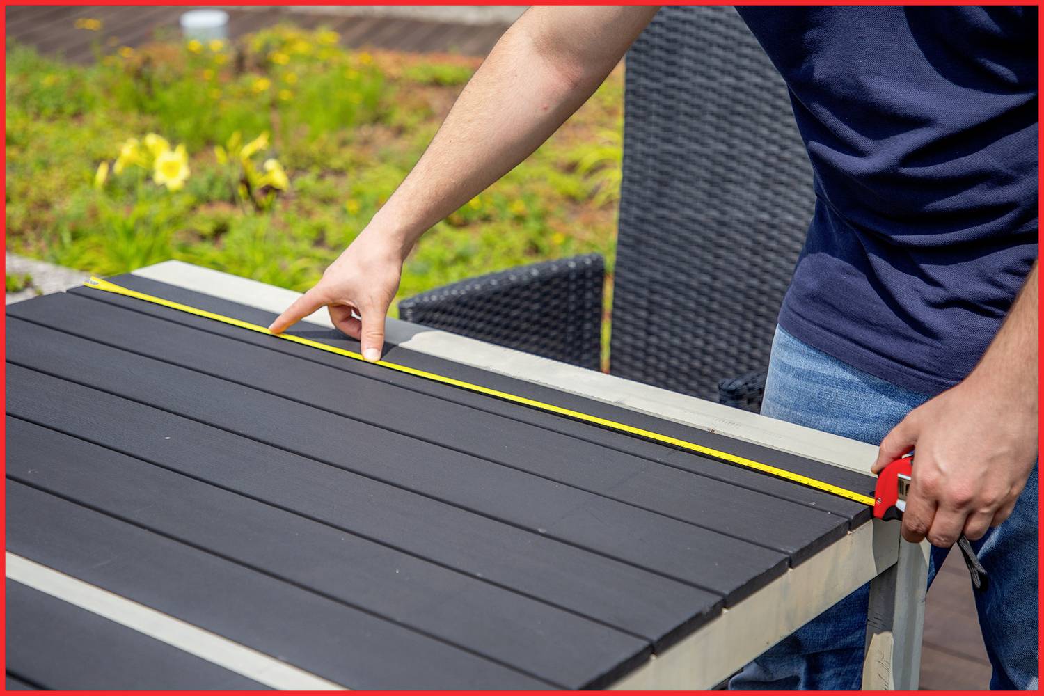 Eine Person misst mit einem Maßband die Länge eines Tisches auf einer Terrasse. Im Hintergrund sind Blumen zu sehen.