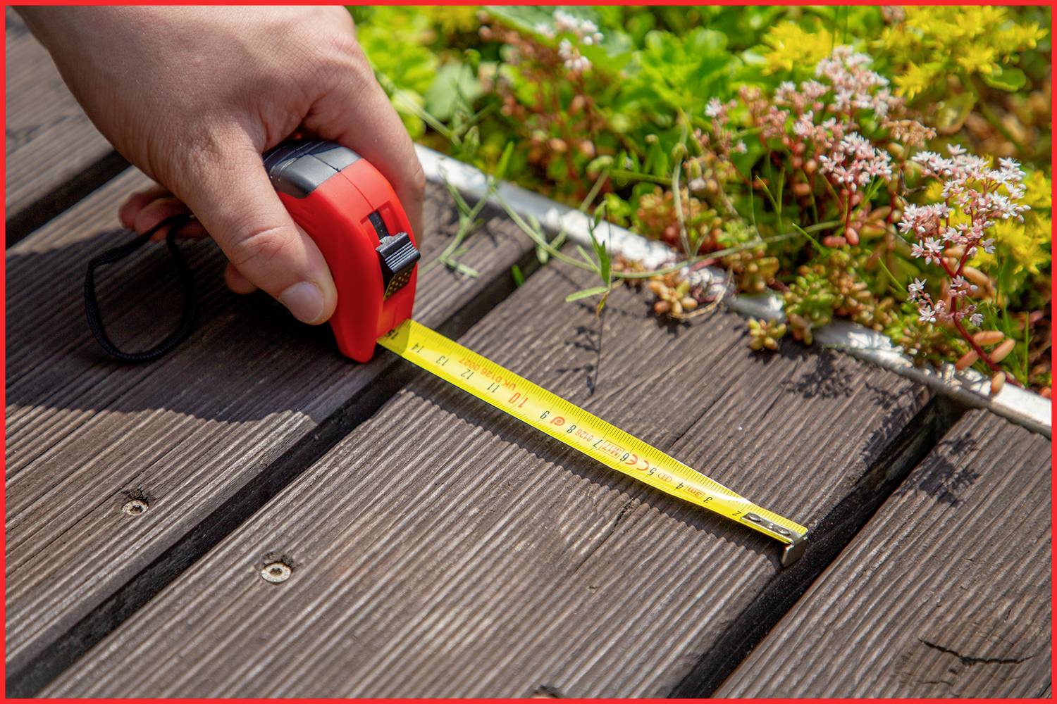 Eine Hand hält ein Maßband auf einer hölzernen Terrasse neben bunten Pflanzen und misst die Länge der Terrassendielen.