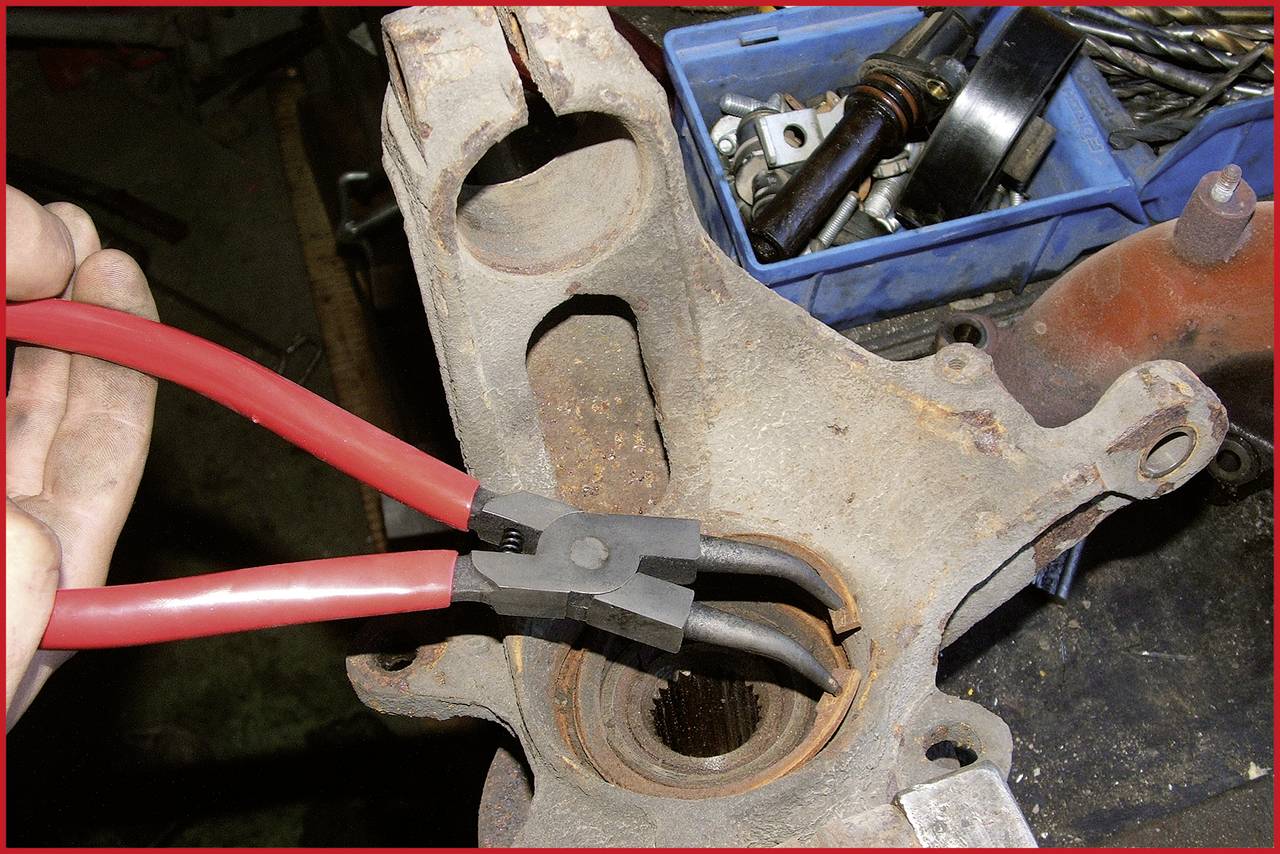 Eine Hand hält eine rote Zange und entfernt sorgfältig einen Sicherungsring von einem Autoteil, das zur Reparatur auf einer Werkbank liegt.