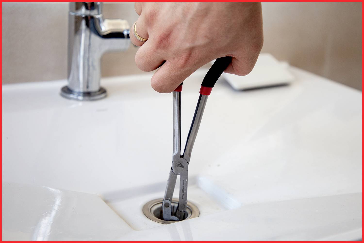 Hand benutzt Zange, um Abfluss in weißem Waschbecken zu reparieren. Nahe Perspektive; Fokus auf Wartung im häuslichen Umfeld.