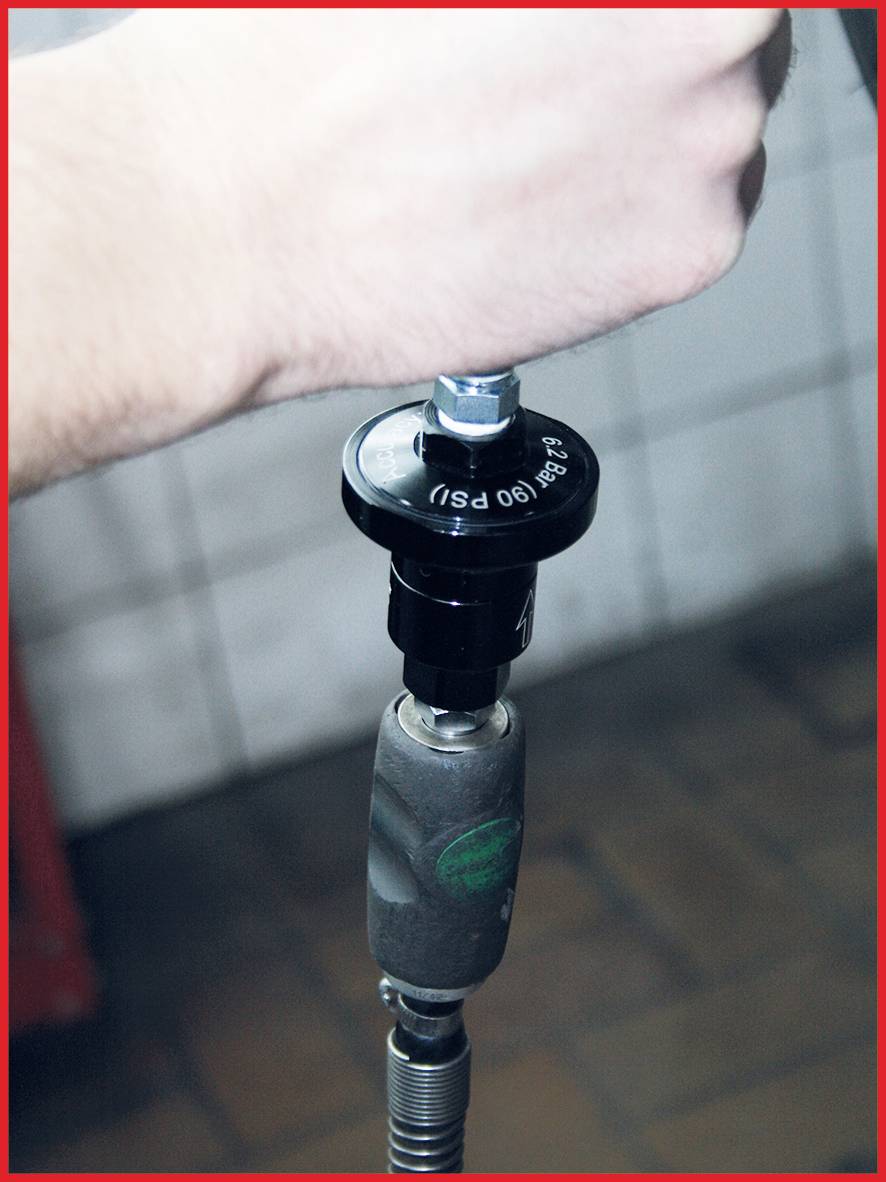 Eine Hand hält ein Werkzeug, das an einem Druckregler befestigt ist. Das Bild zeigt einen technischen Arbeitsprozess in einer Werkstattumgebung.