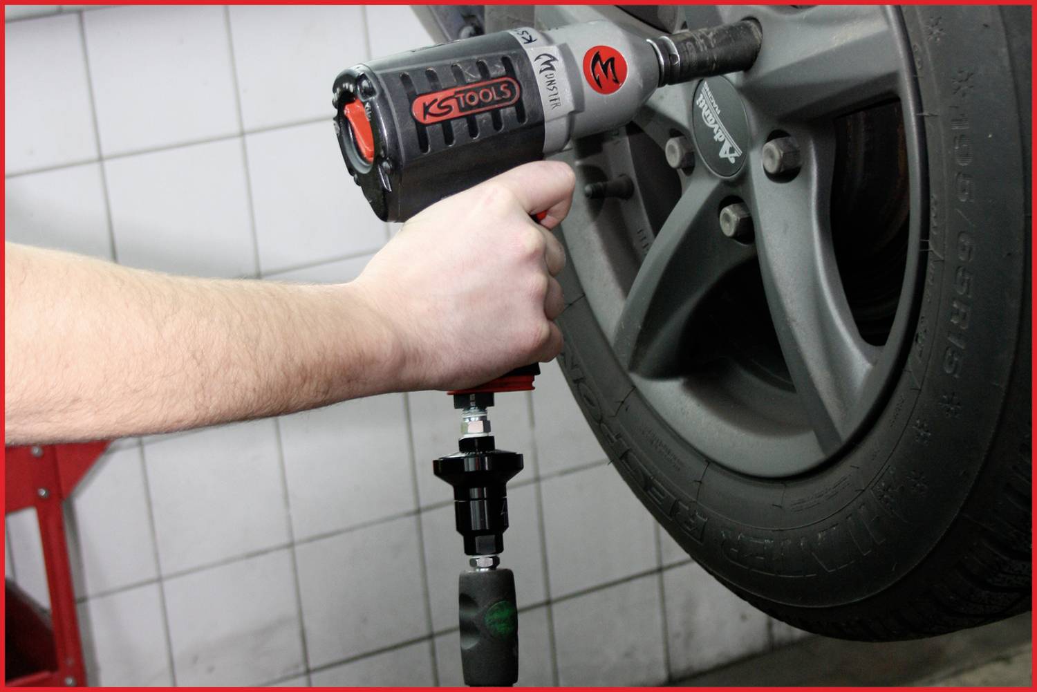 Hand führt Schlagschrauber an Autoreifen an, um Radmontage oder -demontage durchzuführen. Werkstattfliesen im Hintergrund.