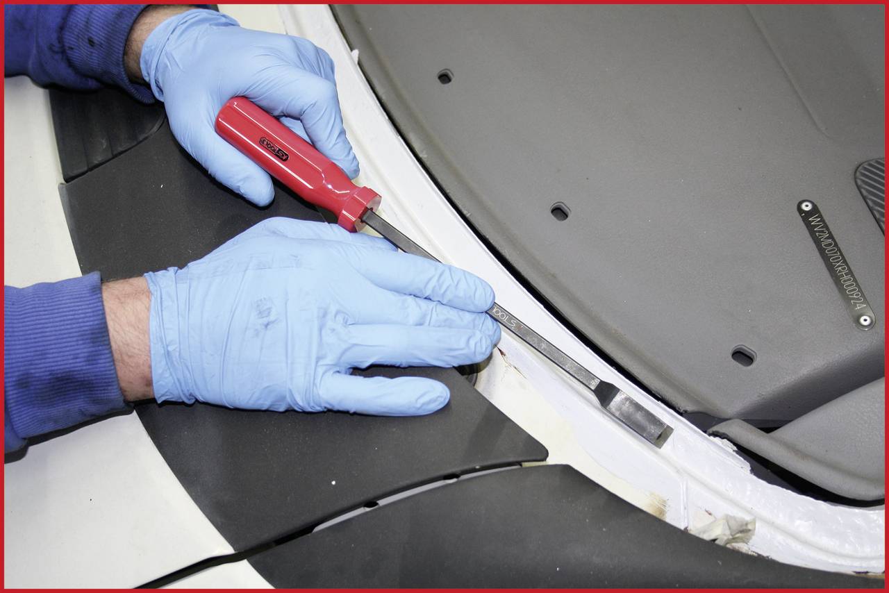 Eine Person trägt blaue Handschuhe und benutzt einen Schraubendreher, um ein Teil an einem Fahrzeug zu lösen.