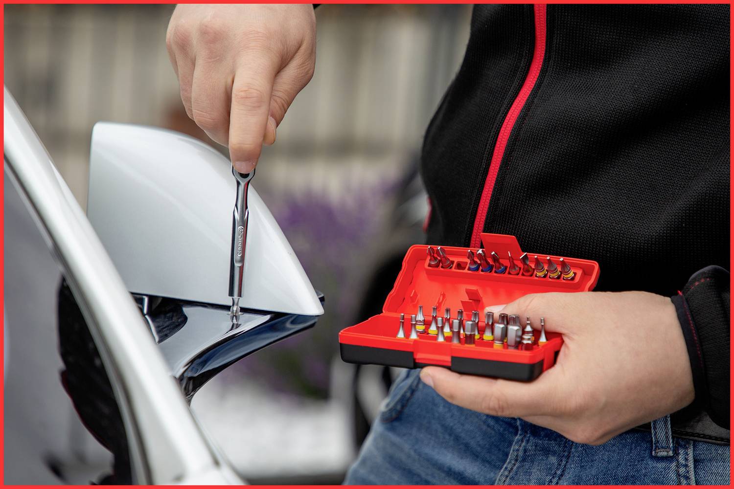 Person hält einen Schraubendreher und ein Set von Schraubendreherbits in der Hand, während sie an einem Außenspiegel eines Autos arbeitet.