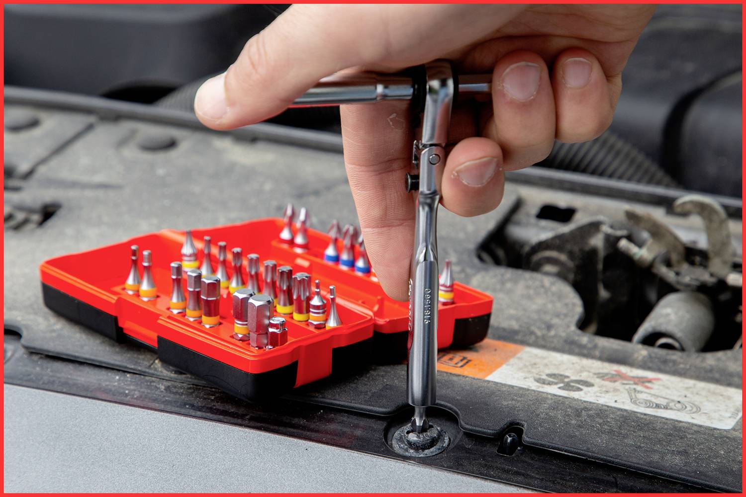 Eine Hand verwendet einen Schraubendreher mit Steckaufsatz, um eine Schraube an einem Auto zu lösen. Im Hintergrund ist ein Set mit verschiedenen Bits.