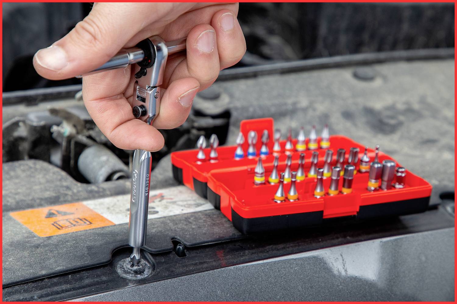 Hand zieht Schraube mit Ratsche in Auto an, nahe offenem Werkzeugkasten mit verschiedenen Aufsätzen.