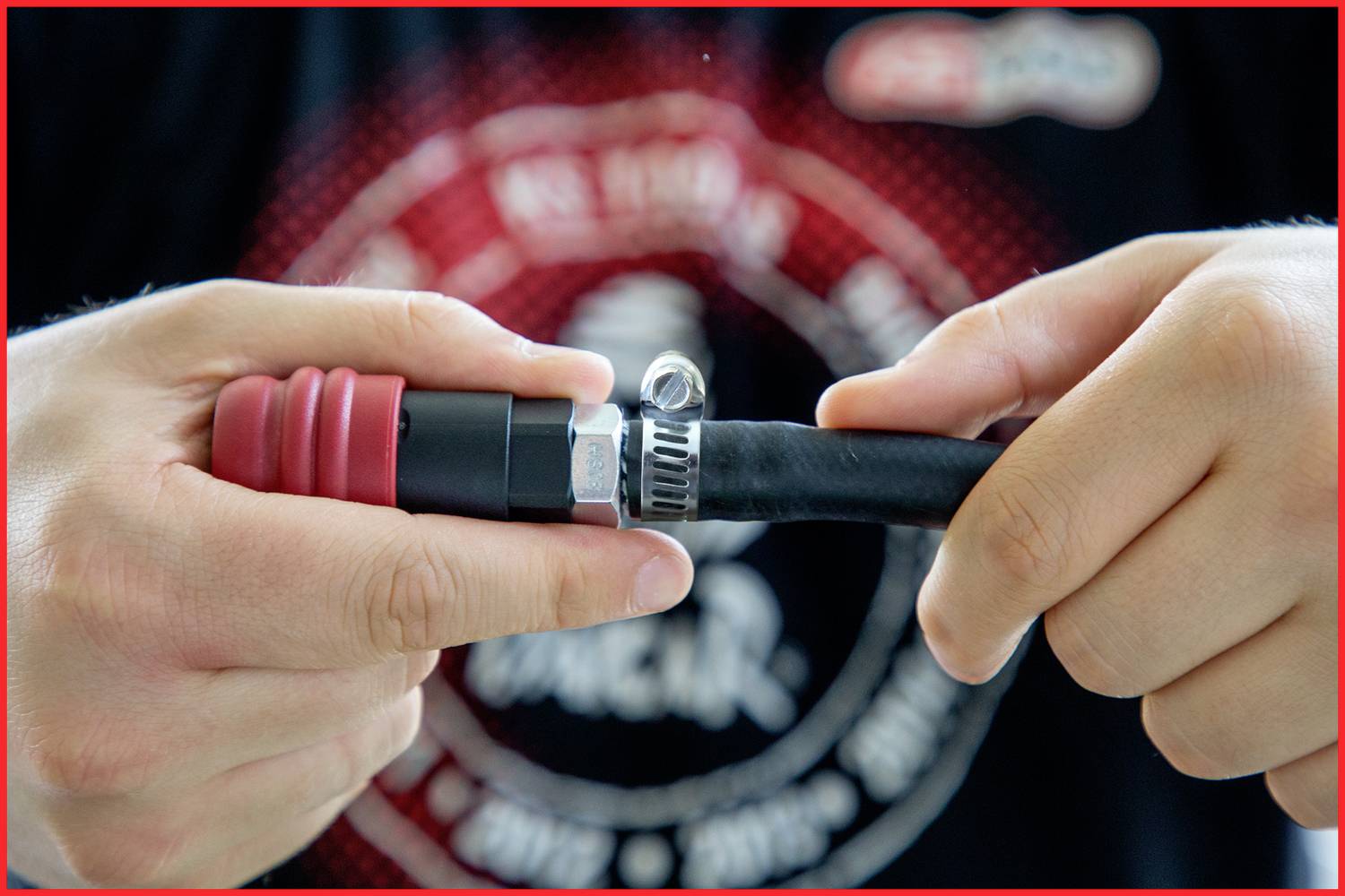 Zwei Hände befestigen einen Schlauch mit einer Schlauchschelle an einem Anschluss. Im Hintergrund ist unscharfe Kleidung zu sehen.