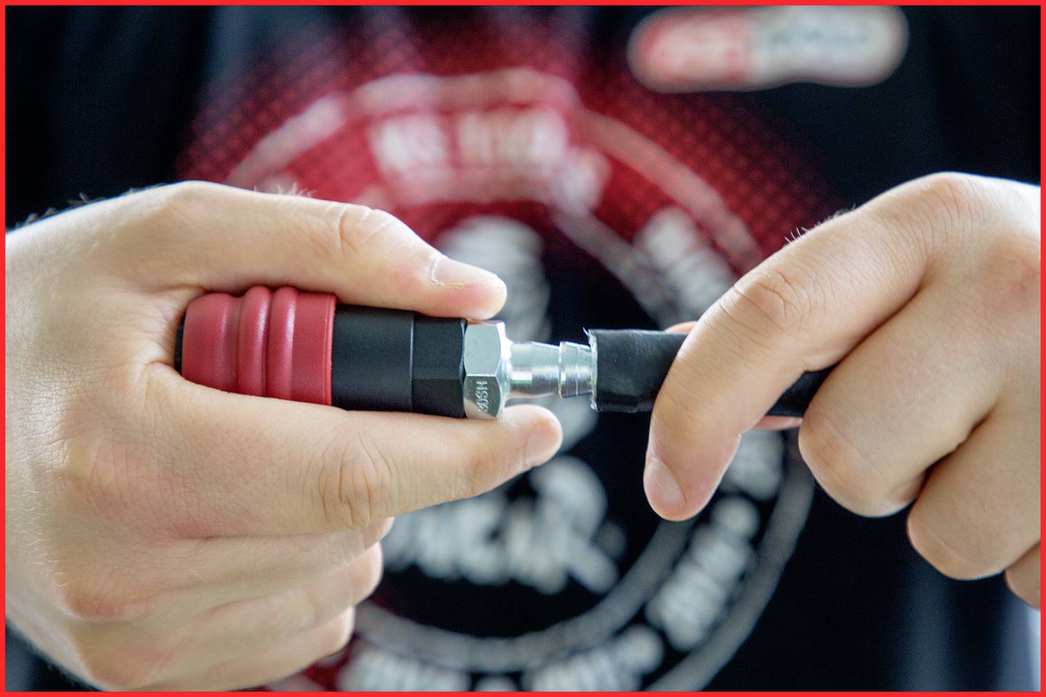 Nahaufnahme von Händen, die einen roten, geriffelten Schlauchanschluss festziehen. Im Hintergrund unscharf ein schwarzes T-Shirt mit Aufschrift.