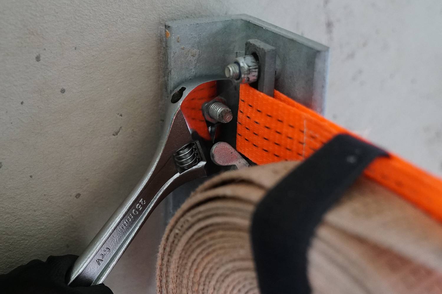 Ein Schraubenschlüssel zieht eine Schraube an, um einen orangefarbenen Spanngurt an einer Metallhalterung zu befestigen. Links ist eine Hand mit Handschuh sichtbar.