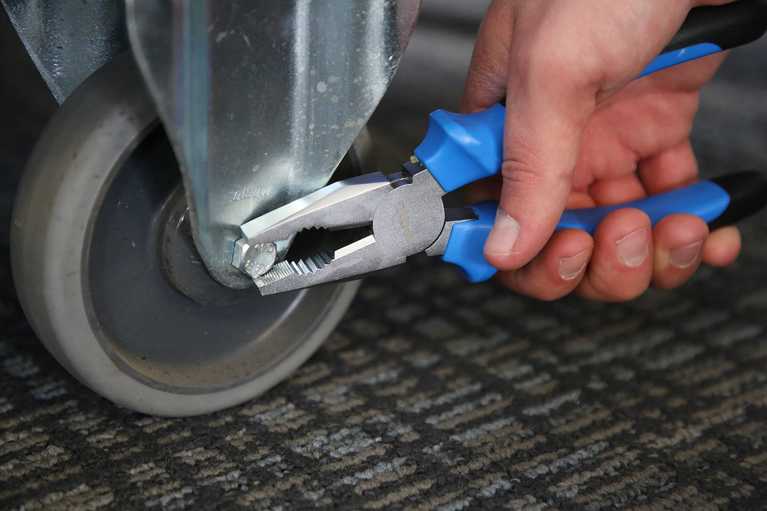 Eine Hand hält eine blaue Zange und zieht die Schraube eines Vorderrades eines Metallwagens an. Der Wagen steht auf einem Teppichboden.