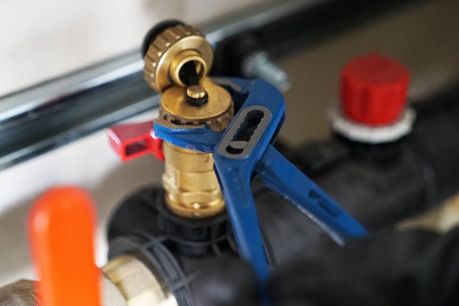 Eine blaue Rohrzange hält ein Messingventil in einem Rohrsystem. Das Ventil hat einen roten Hebel. Im Hintergrund sind Teile der Rohrinstallation zu sehen.