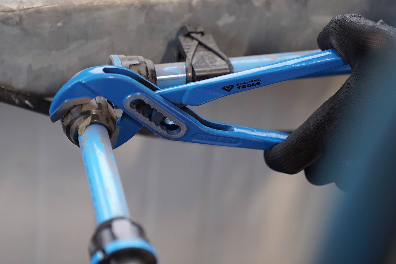 Eine Person zieht mit einer blauen Rohrzange an einem Rohr. Die Hand trägt einen schwarzen Handschuh, Hintergrund unscharf.