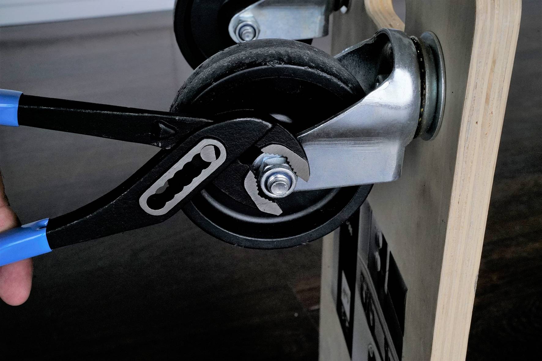 Eine schwarze Zange hält die Achse eines schwarzen Radcastors, der an einem Holzbrett befestigt ist. Dieses wird anscheinend repariert oder justiert.