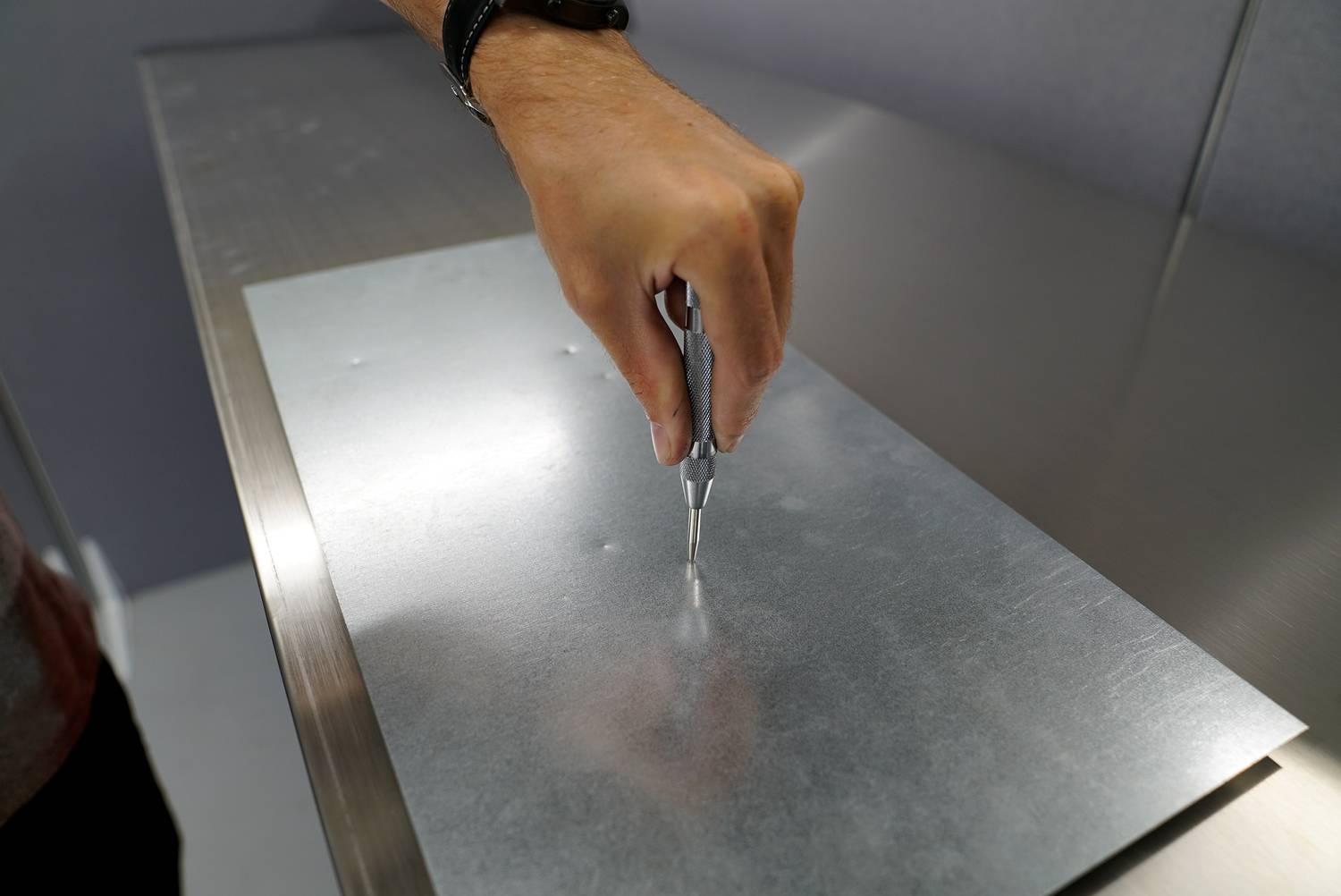 Eine Hand hält ein Werkzeug über einem Metallblech und setzt einen Punkt. Die Szene zeigt einen Arbeitsschritt in der Metallbearbeitung.