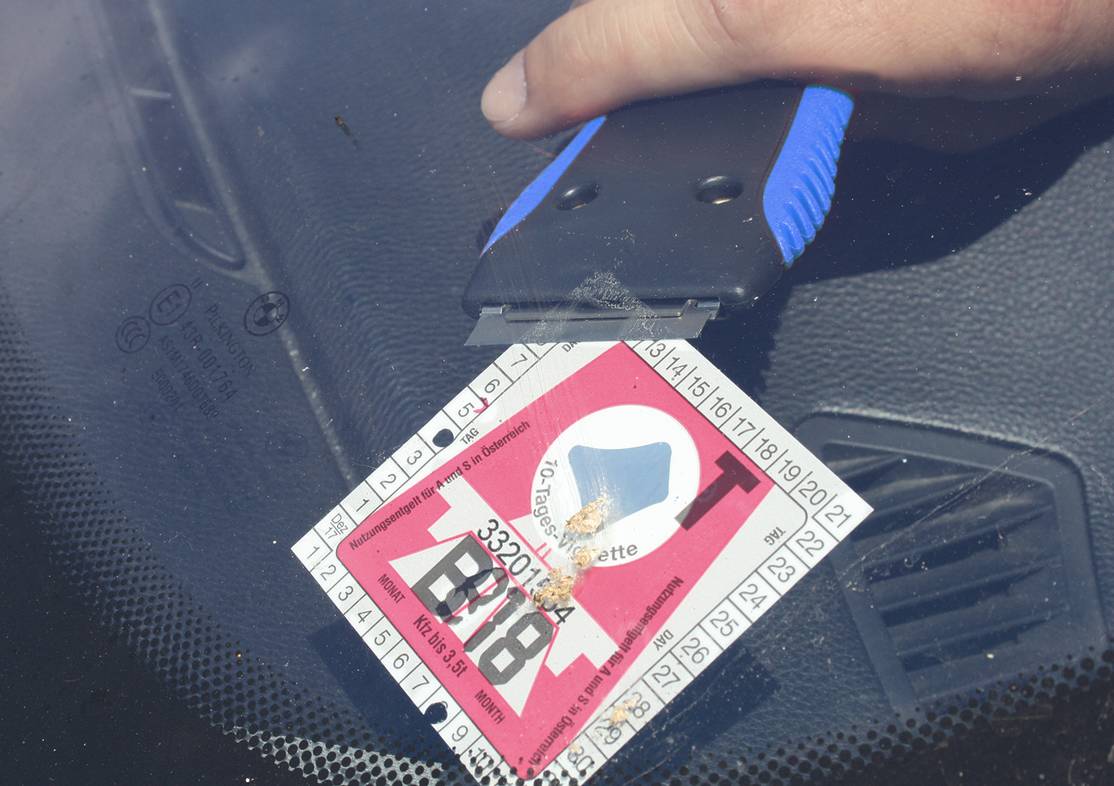 Eine Hand entfernt mit einem Schaber einen österreichischen Autobahn-Vinjettenaufkleber von einer Windschutzscheibe.