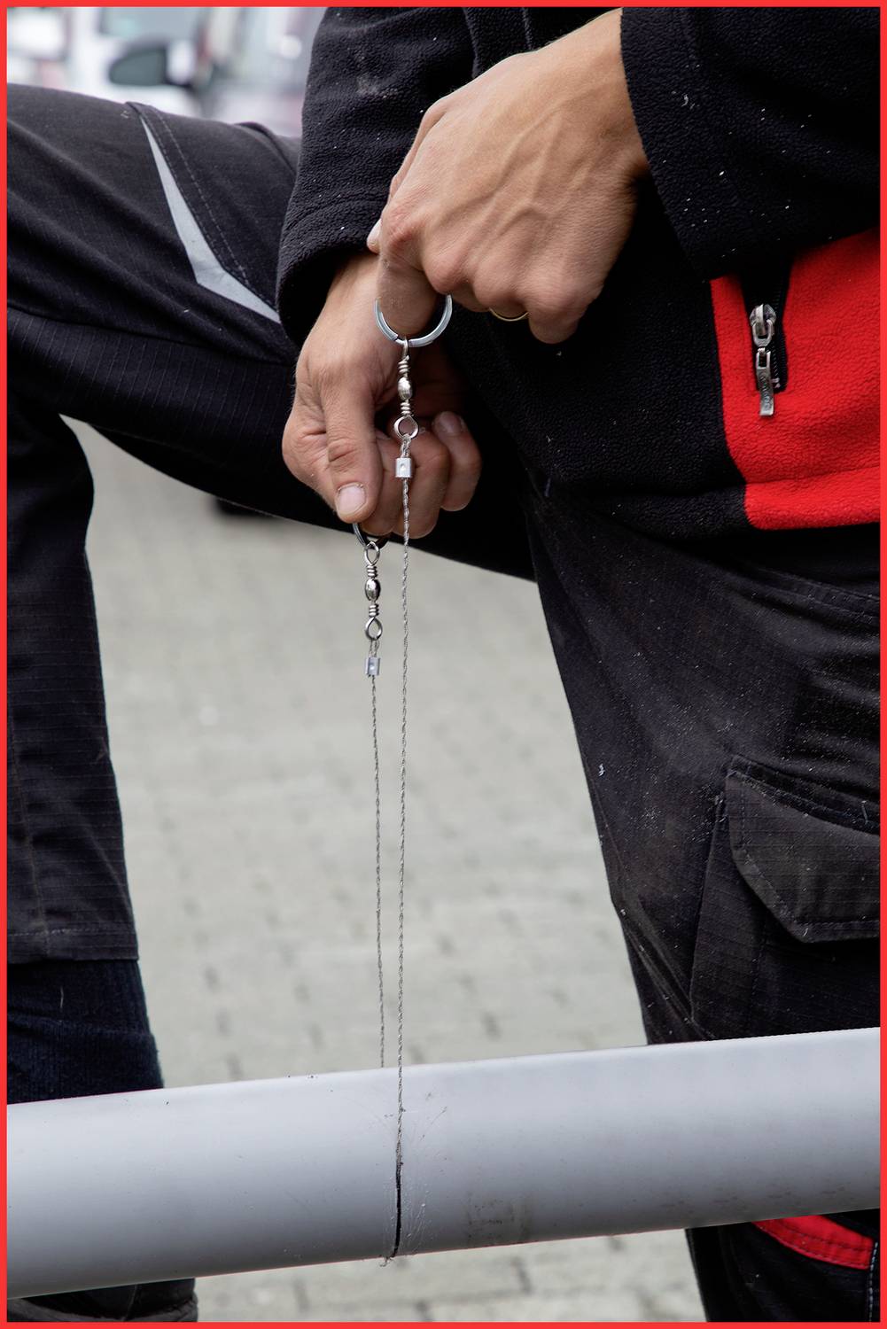 Eine Person im schwarzen und roten Outfit dreht ein Metallteil in der Hand. Der Hintergrund zeigt eine gepflasterte Fläche.