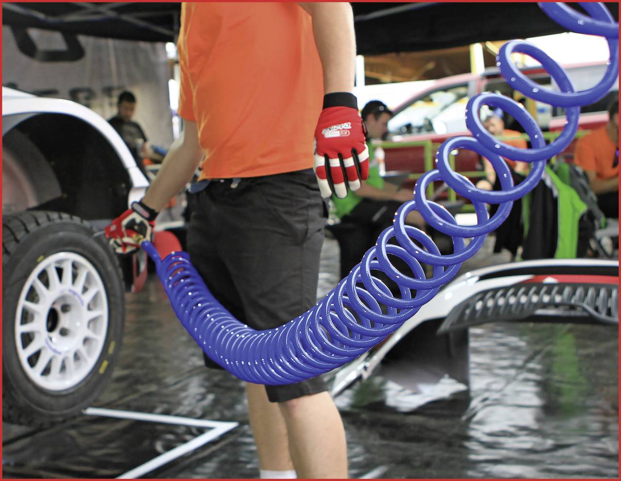 Eine Person in einem orangefarbenen Hemd hält einen langen, blauen Luftschlauch vor einem Auto in einer Werkstattumgebung.