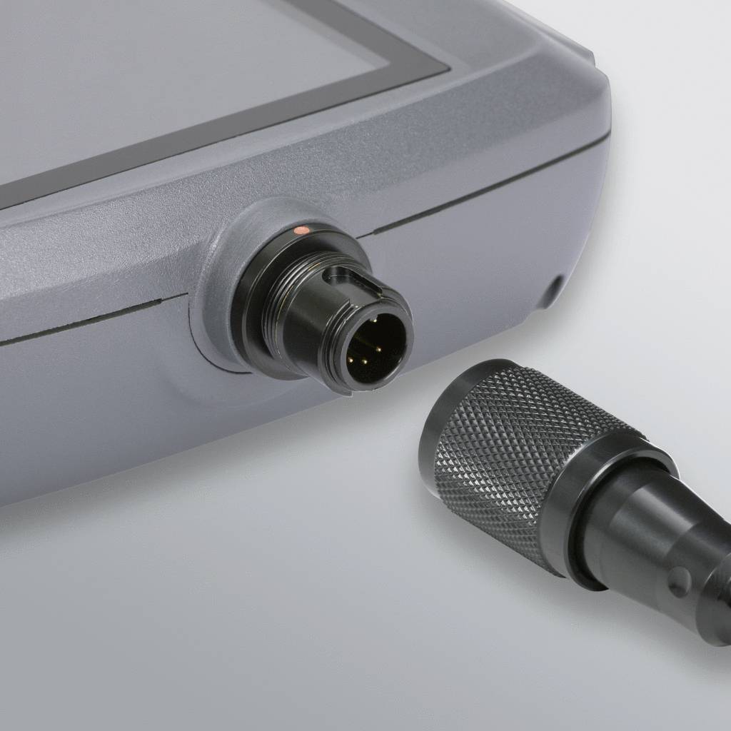 Ein graues Gerät mit einem runden Anschluss, neben dem ein abnehmbarer Stecker liegt, zeigt eine Verbindungssituation.