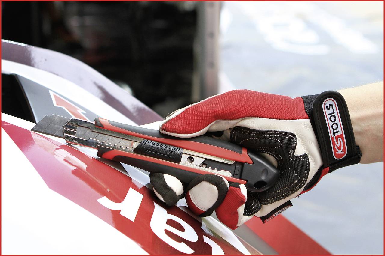 Hand mit Handschuh schneidet mit einem Cuttermesser einen roten Aufkleber auf einem Auto. Werkzeugmarke deutlich erkennbar.