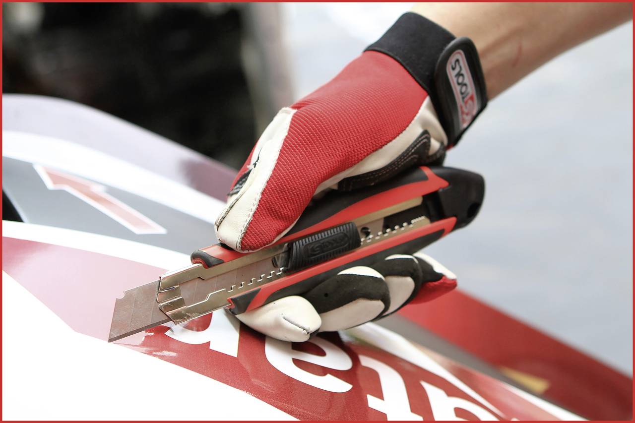 Hand in roten Handschuhen hält ein Cuttermesser, das auf einem rot-weißen Untergrund positioniert ist.